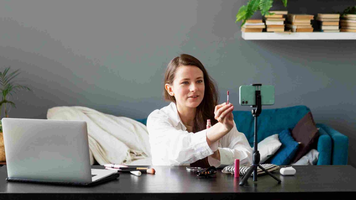 A woman creating a beauty vlog using her smartphone, sitting at a table with makeup products and a ring light in a cozy setting.