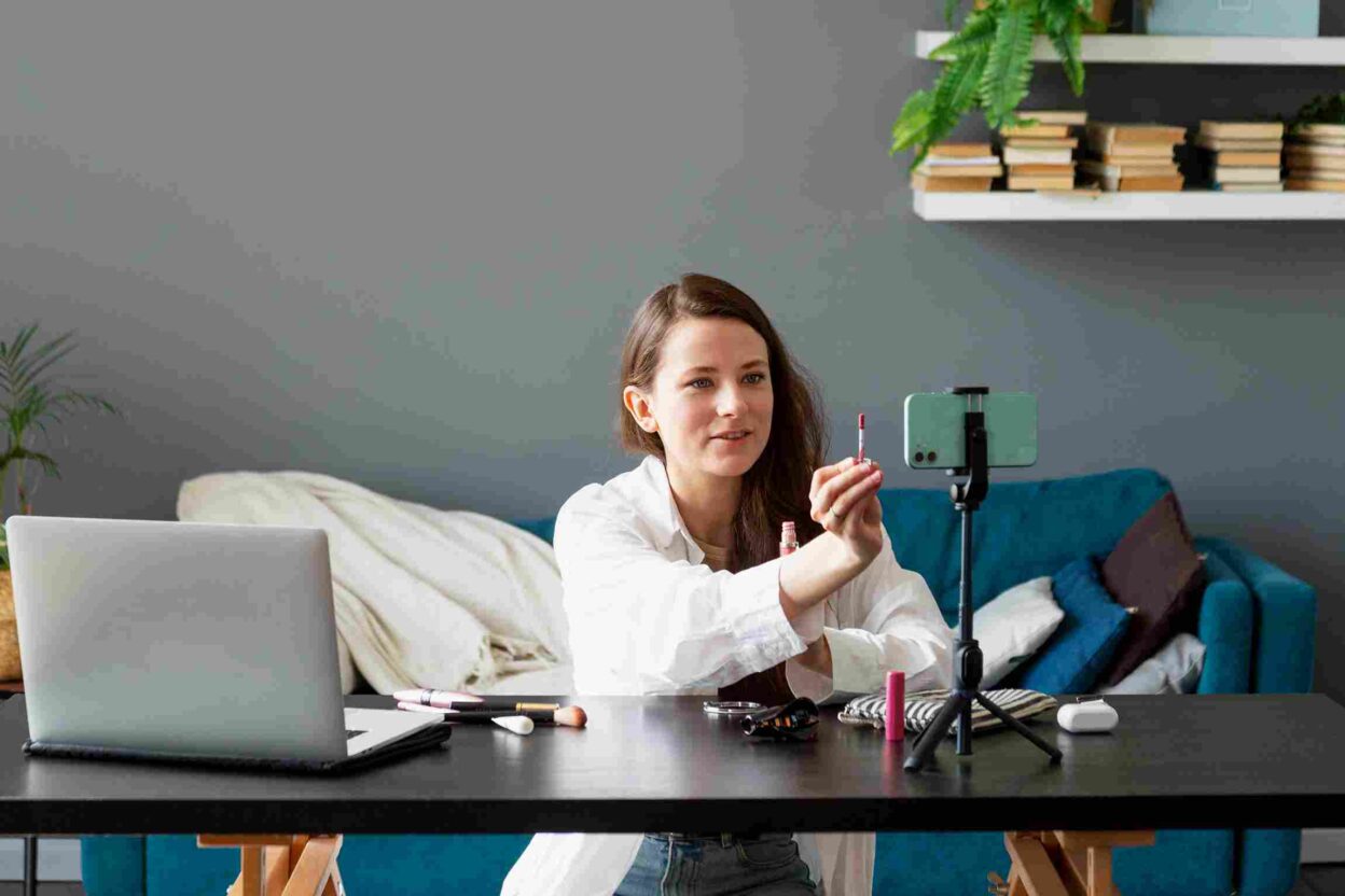 A woman creating a beauty vlog using her smartphone, sitting at a table with makeup products and a ring light in a cozy setting.